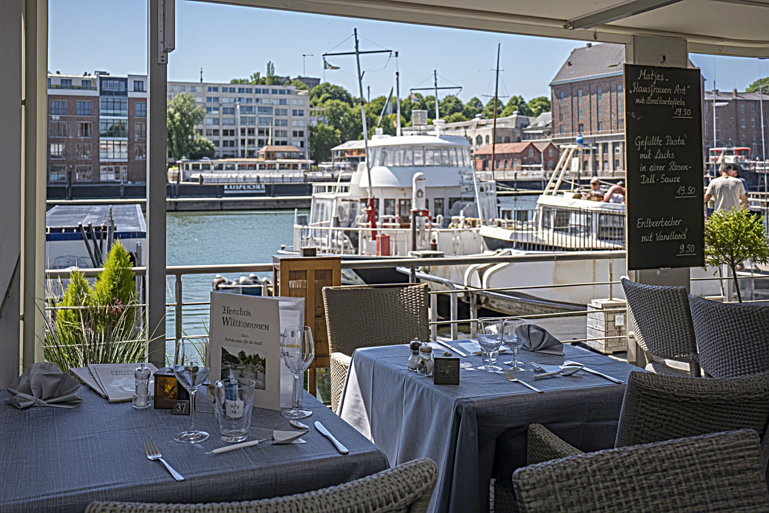 Sonnige Restaurantterrasse am Hafen mit gedeckten Tischen und Blick aufs Wasser